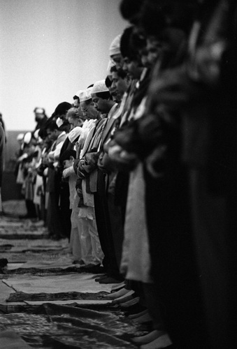 A group of men and boys standing in rows, facing forward, with their hands clasped in front of them during prayer.