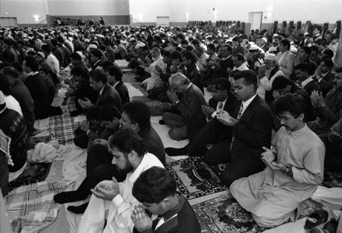 A large group of men sitting on prayer mats, many with their hands open in prayer, looking towards the front.